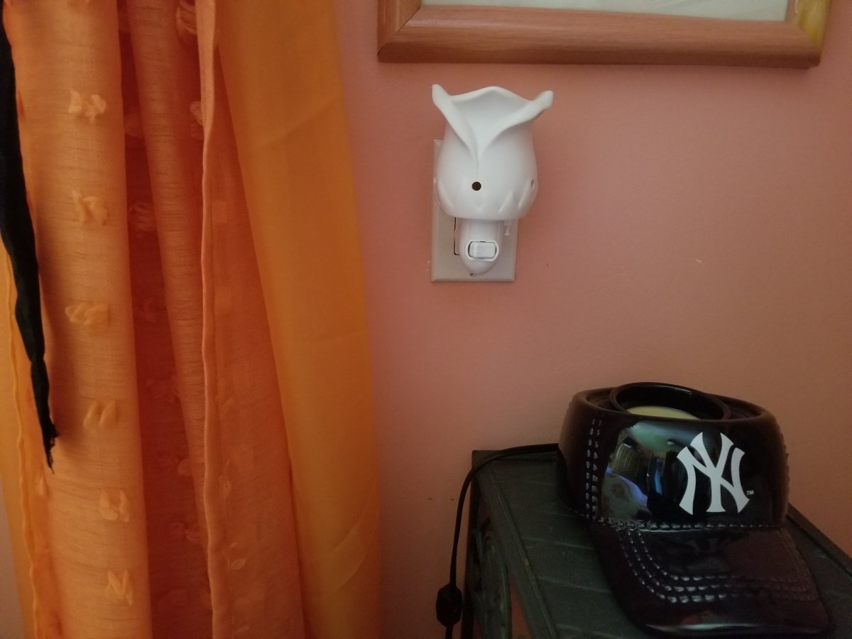A rose shaped white wax warmer is lit up and plugged into a wall. Below it is another wax warmer in the shape of a baseball helmit with the New York Yankees logo on the helmit