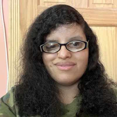 A woman with long, curly black hair and wearing glasses is smiling at the camera. She is dressed in a camouflage-patterned top. The background features a wooden door and a wall painted in a light peach color.