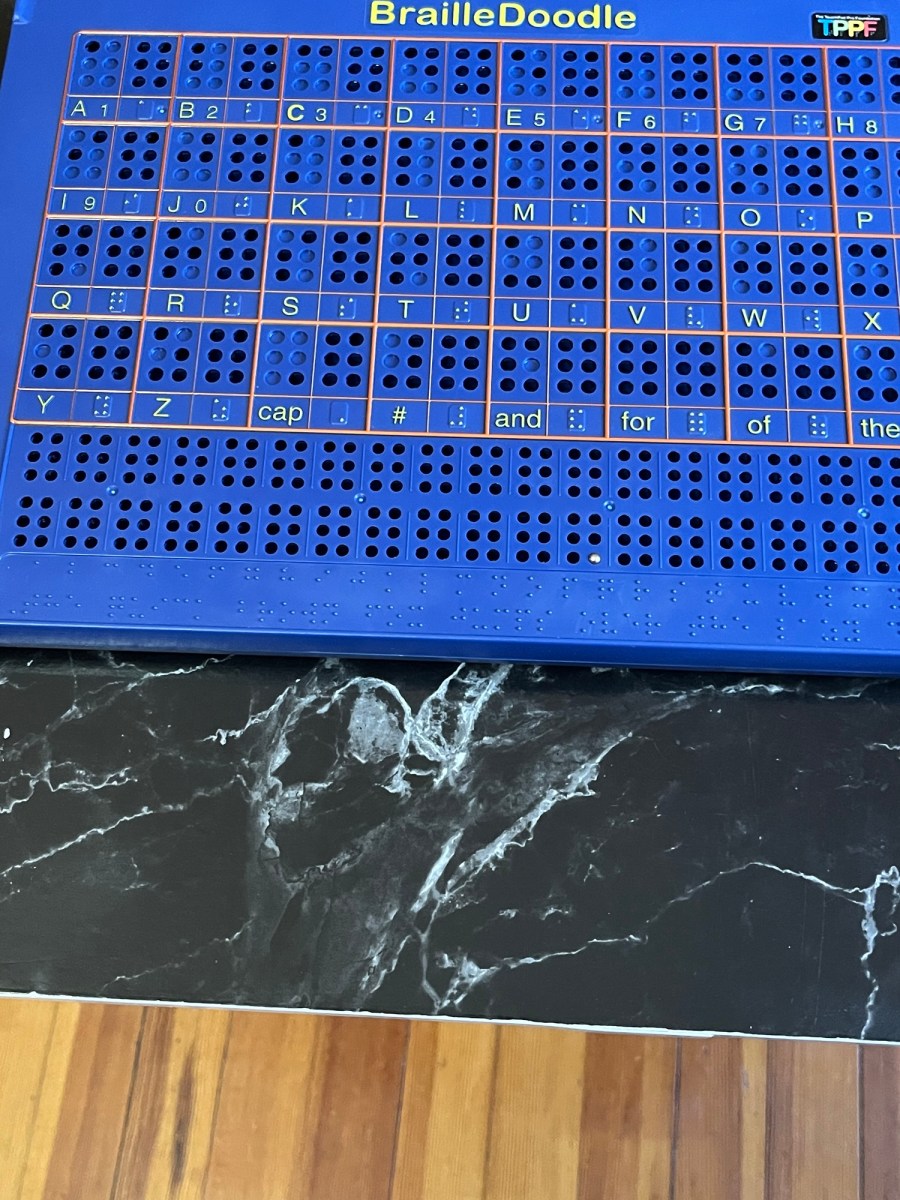 Blue Braille learning board on a table. The board is labeled "BrailleDoodle" at the top and has a grid with raised Braille dots for each letter of the alphabet (A-Z), numbers 1 to 0, and common Braille contractions like "cap," "#," "and," "for," "of," and "the." Each cell in the grid includes both the Braille character and its printed equivalent. Below the main grid, there are several blank rows with only Braille dots, which might be for practice. The board is resting on a black surface with white marble patterns, and below that, a wood floor is visible.