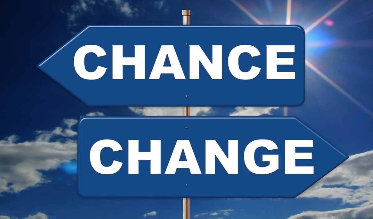 Two blue directional signs with white capital letters, one on top of the other, against a sky with clouds and sun rays. The top sign points left and says "CHANCE". The bottom sign points right and says "CHANGE". The signs are mounted on the same pole.