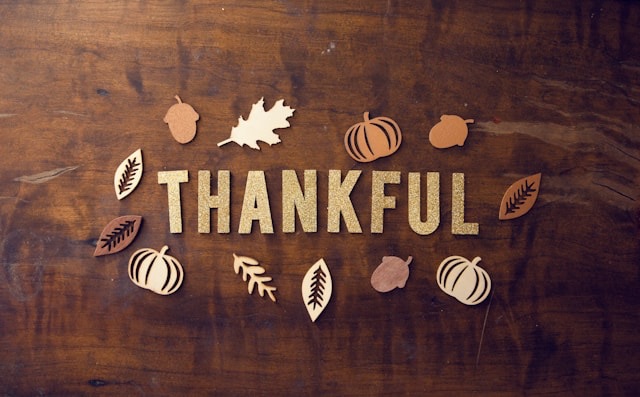 Wooden table surface with the word "THANKFUL" in large, sparkly gold letters at the center. Surrounding the word are small wooden cutouts of autumn leaves, acorns, and pumpkins in brown, orange, and cream colors, arranged in a scattered pattern for a festive, autumnal look.